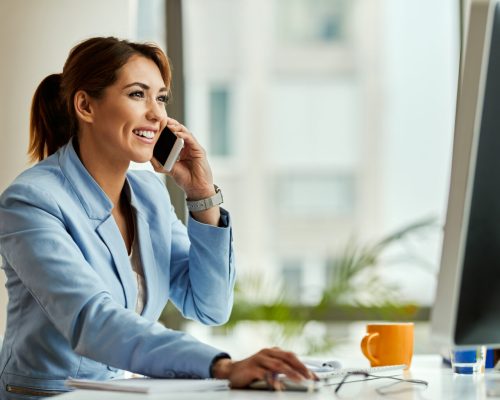 Young happy businesswoman working on desktop PC and communicating over mobile phone in the office.